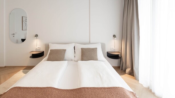 Ein modernes Schlafzimmer mit einem großen Bett, zwei Nachttischen mit Lampen, einem runden Spiegel und Ausblick durch leichte Vorhänge.