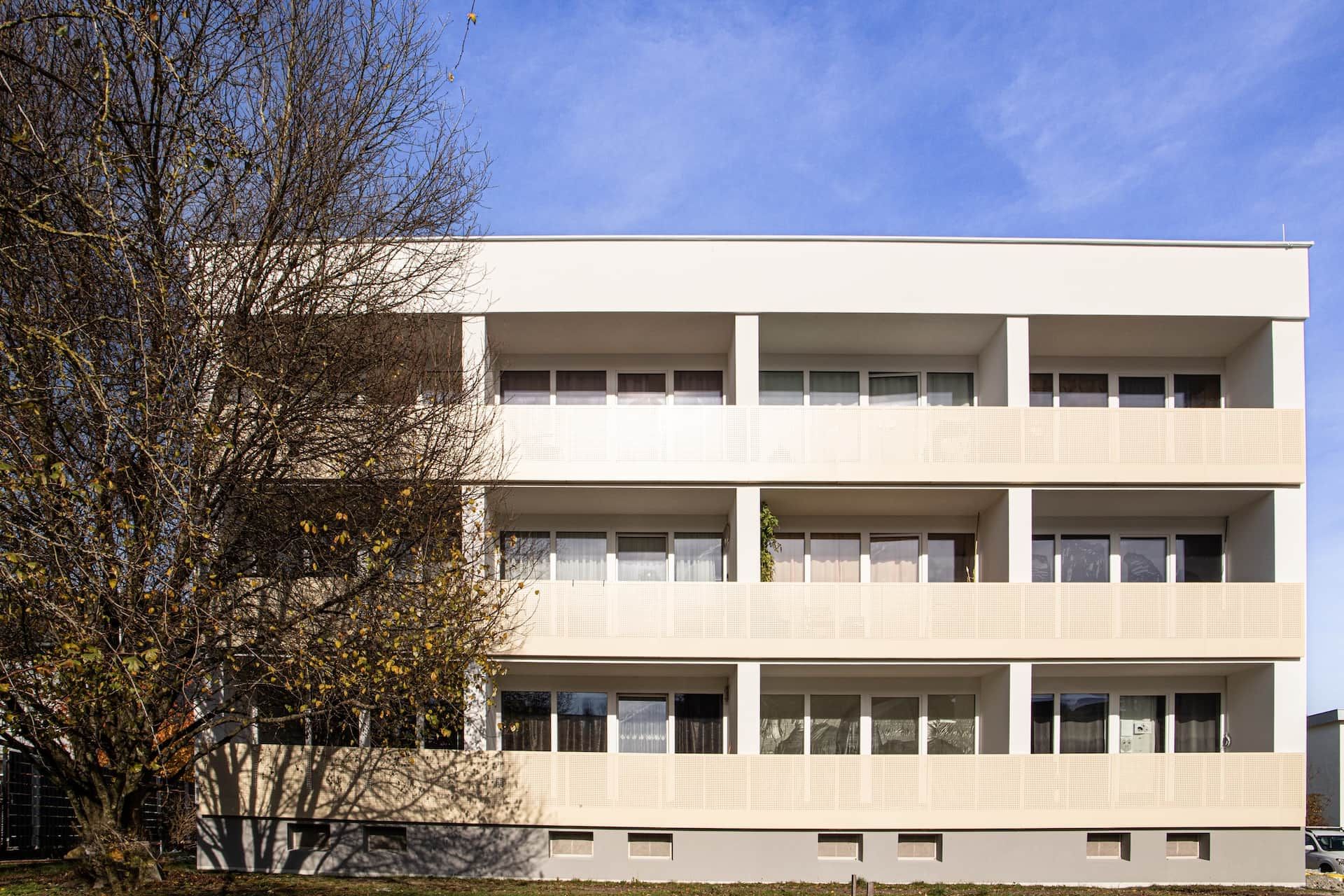 A multi-story building with a flat facade, large windows, and a tree in the foreground.