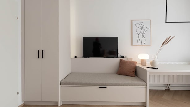 A modern room with a built-in bench, a flat-screen TV, a framed abstract art piece, and a desk with a lamp.
