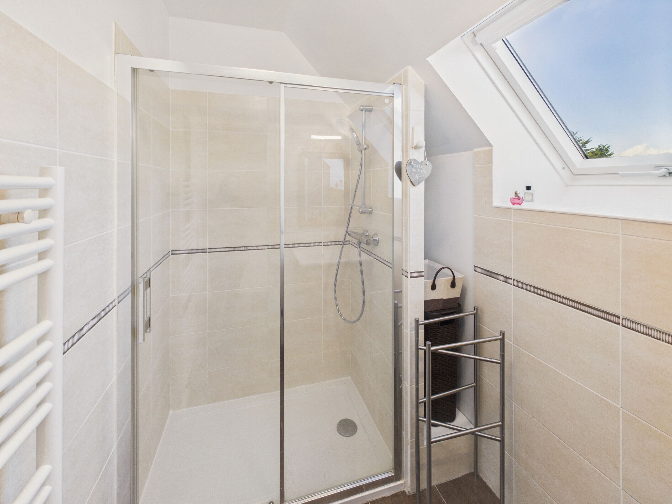 Et brusebad med glasvægge, en håndbruser, et hvidt håndklædetørrestativ og en lille hylde med toiletartikler.