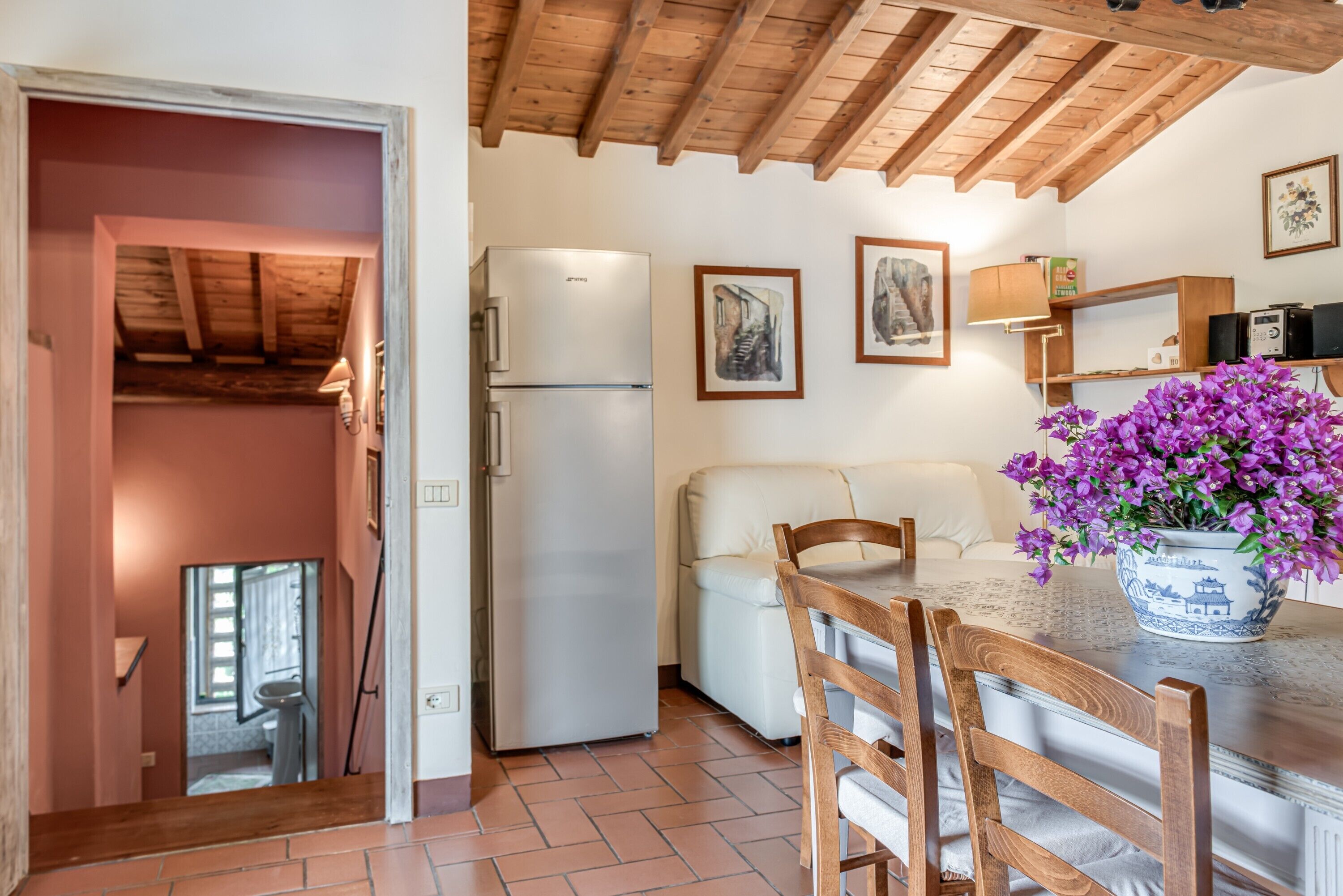 Une cuisine équipée d’un réfrigérateur, avec des poutres en bois au plafond, une table à manger avec des chaises et un vase rempli de fleurs violettes.