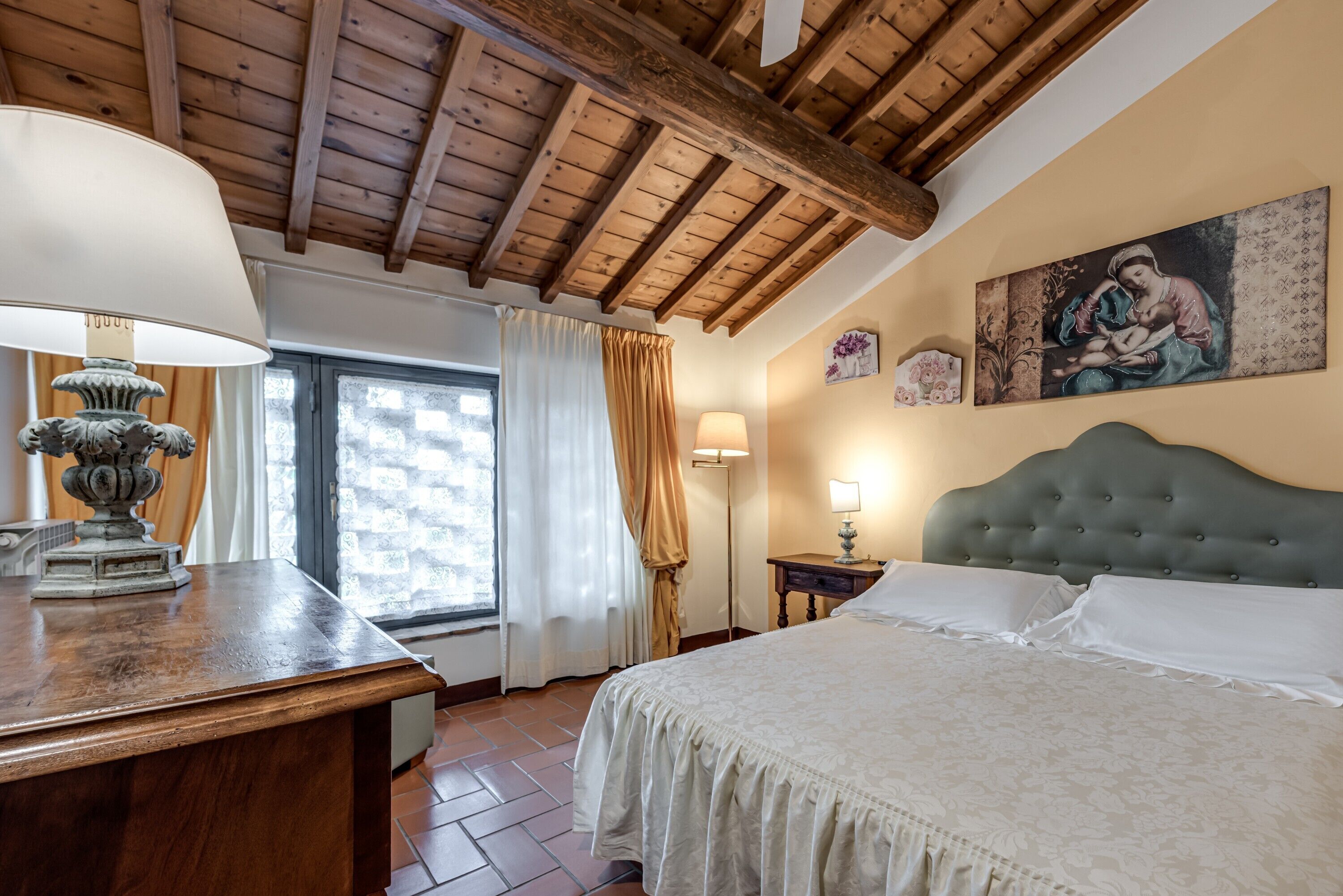 A bedroom with a wooden ceiling, a bed with a grey headboard, a bedside table with a lamp, and a window with curtains.