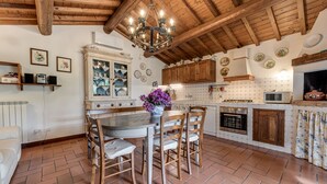Een keuken met houten balken, een kroonluchter, een eettafel met stoelen en een betegelde achterwand.