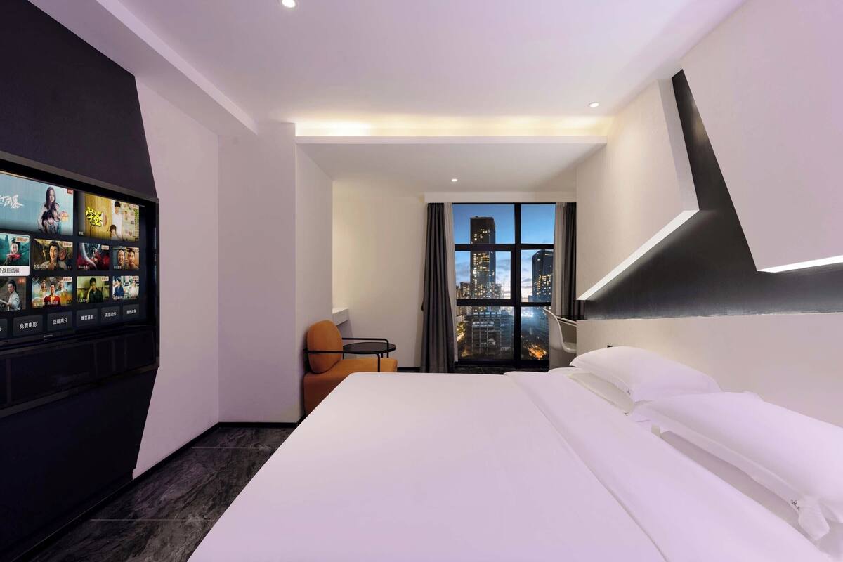 Quarto de hotel com uma cama, uma TV exibindo cartazes de filmes, vista para a cidade pela janela e uma pequena mesa com uma cadeira.