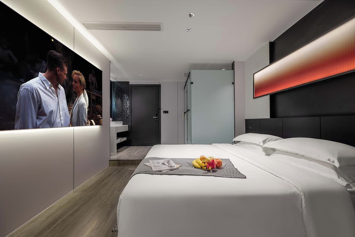Quarto de hotel moderno com uma cama grande, mesa de cabeceira com uma cesta de frutas, televisão fixada na parede e design minimalista.