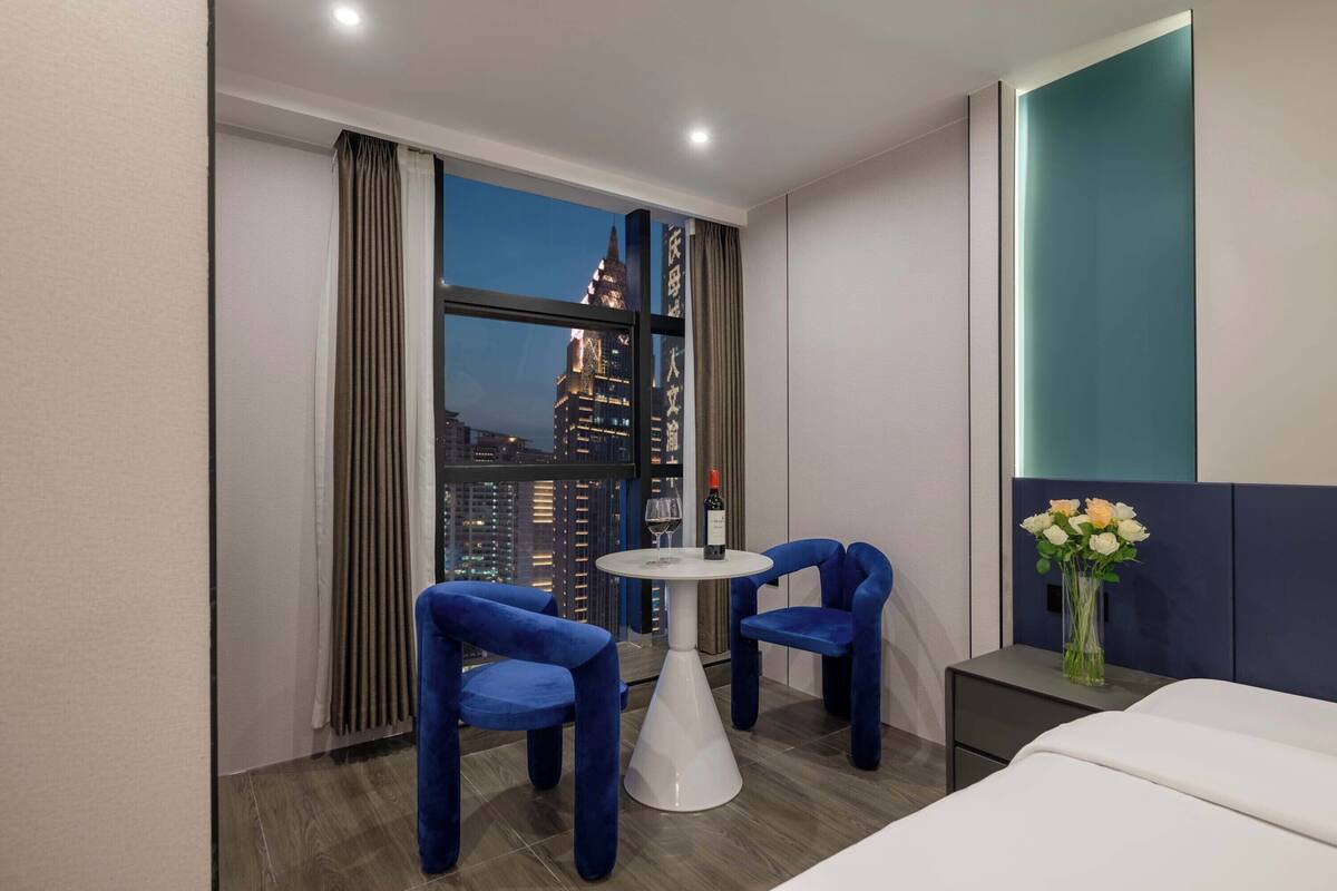Quarto de hotel com vista para a cidade, uma mesa redonda com cadeiras, uma cama e uma mesinha de cabeceira com um vaso de flores.
