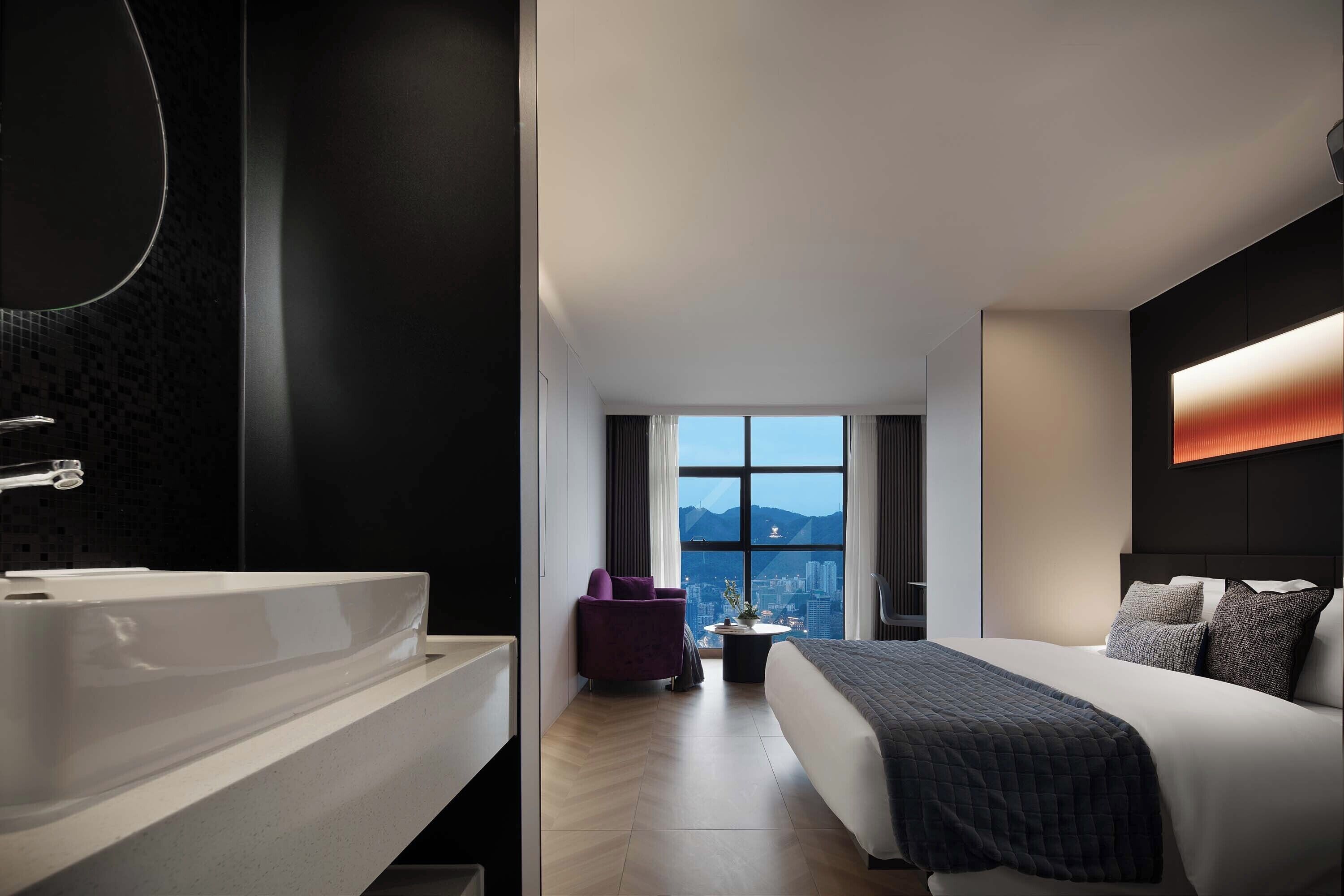 A modern hotel room with a large bed, a bathroom with a bathtub, a city view through the window, and a small seating area.