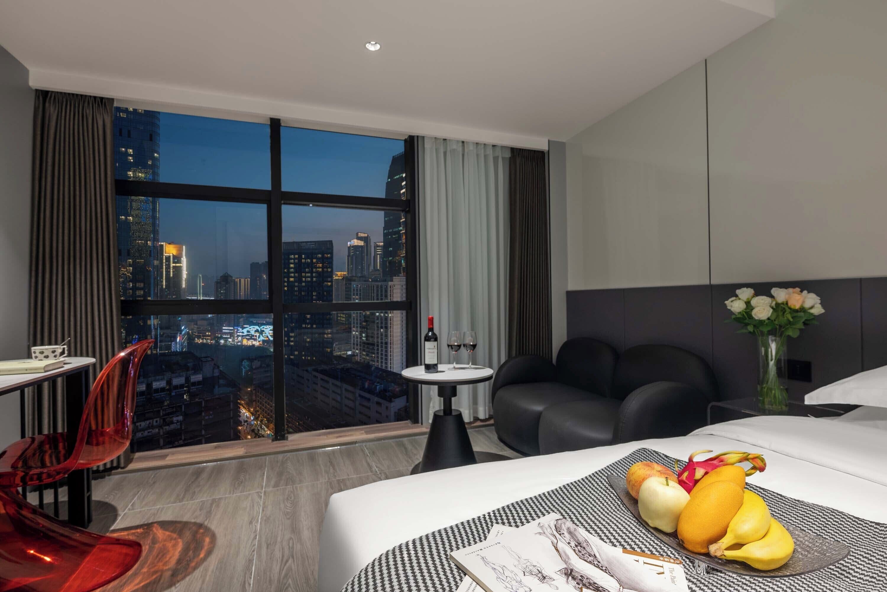 A hotel room with a large window, a dining table, a sofa, and a bed with fruit on the side.