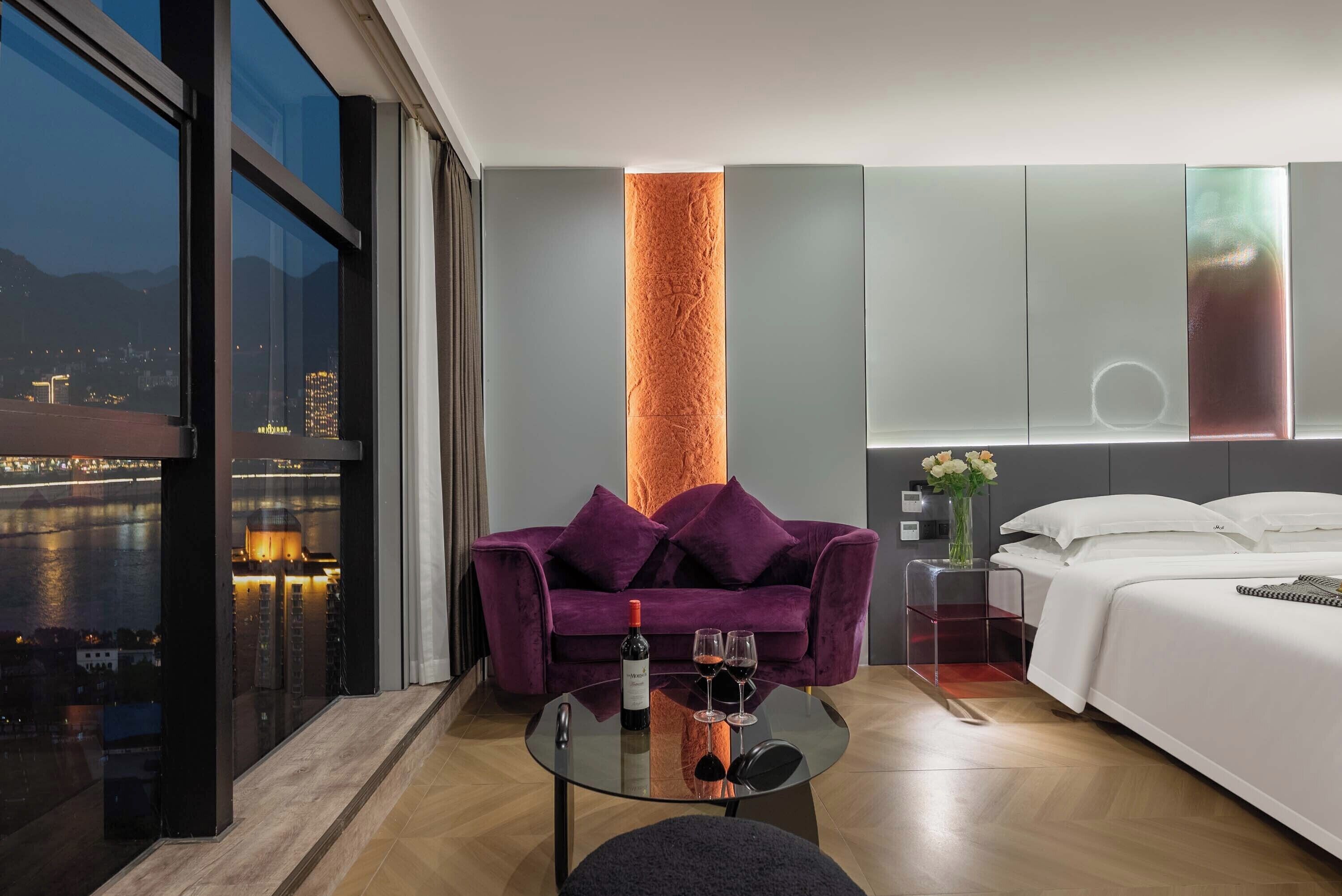 A modern hotel room with a large window, a purple sofa, a glass table with wine glasses, and a bed with white linens.