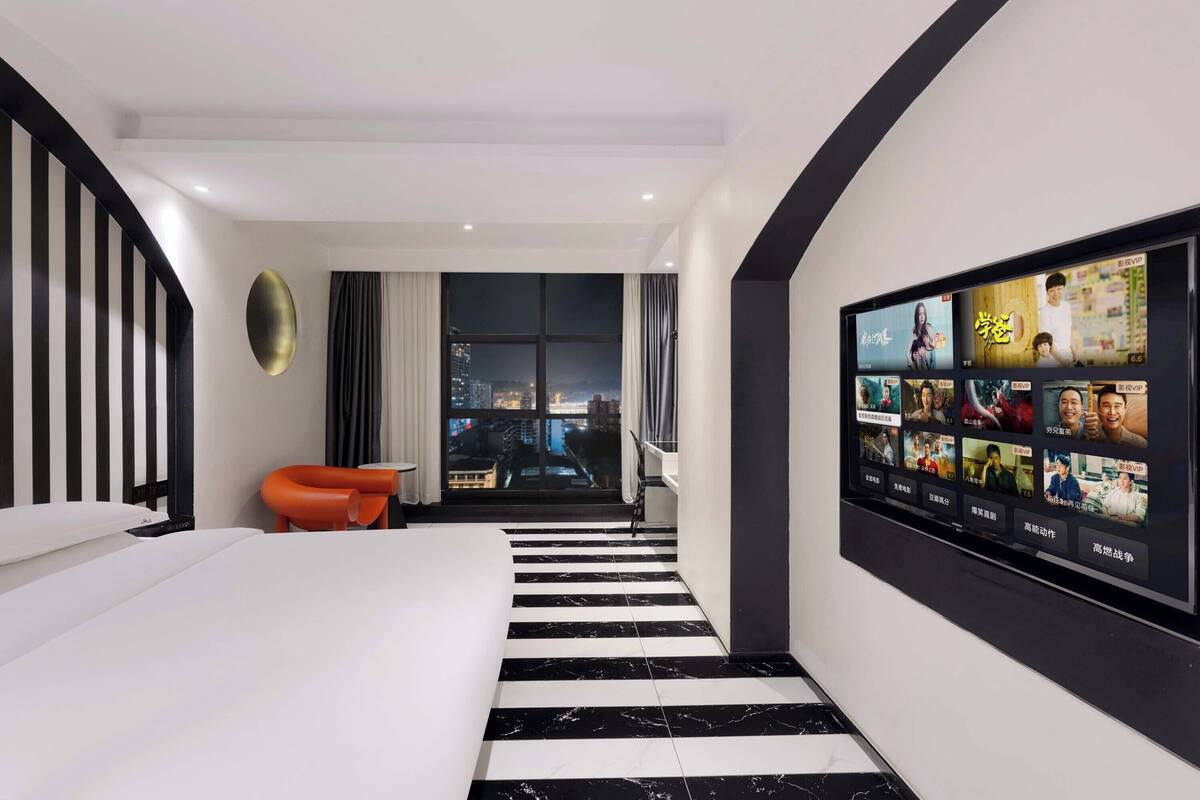Quarto de hotel moderno com uma cama grande, uma TV fixada na parede, vista da cidade por uma grande janela e uma escada que leva a um andar superior.