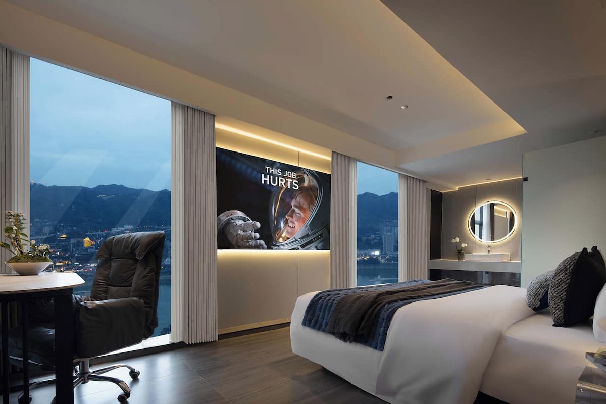 Quarto de hotel moderno com uma cama grande, uma mesa de trabalho e uma cadeira. Há uma janela ampla com vista para a paisagem urbana e montanhas.