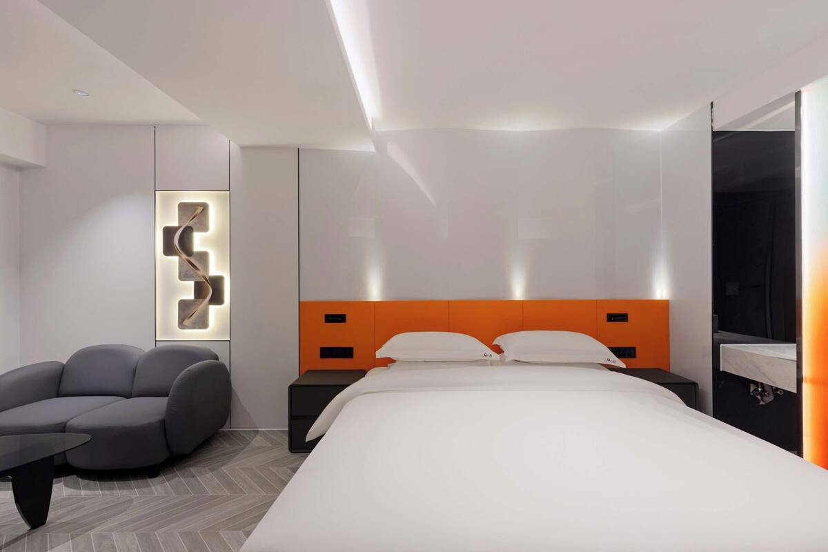 Quarto de hotel moderno com uma cama grande, um sofá cinzento e uma mesa de cabeceira.