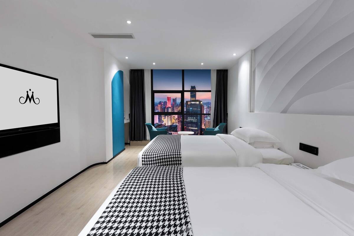 Quarto de hotel moderno com uma janela grande que oferece vista para a cidade, uma cama com roupa de cama branca, um banco estampado em xadrez de cães e uma TV fixada na parede.