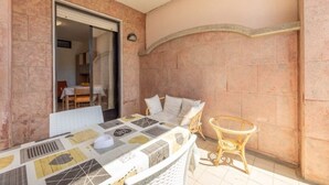 Une terrasse ensoleillée avec une table dressée pour le repas, des chaises en osier et une vue sur l’intérieur à travers une porte vitrée.