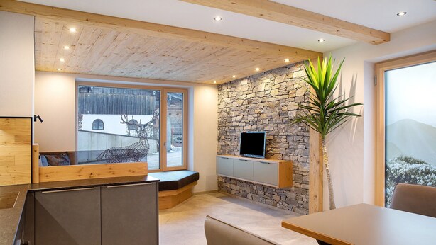A modern interior with a stone wall, a TV mounted on it, a wooden ceiling, and a large window. - Apartment "Wildschön" with Mountain Views, Shared Garden and Wi-Fi (Grafenweg)