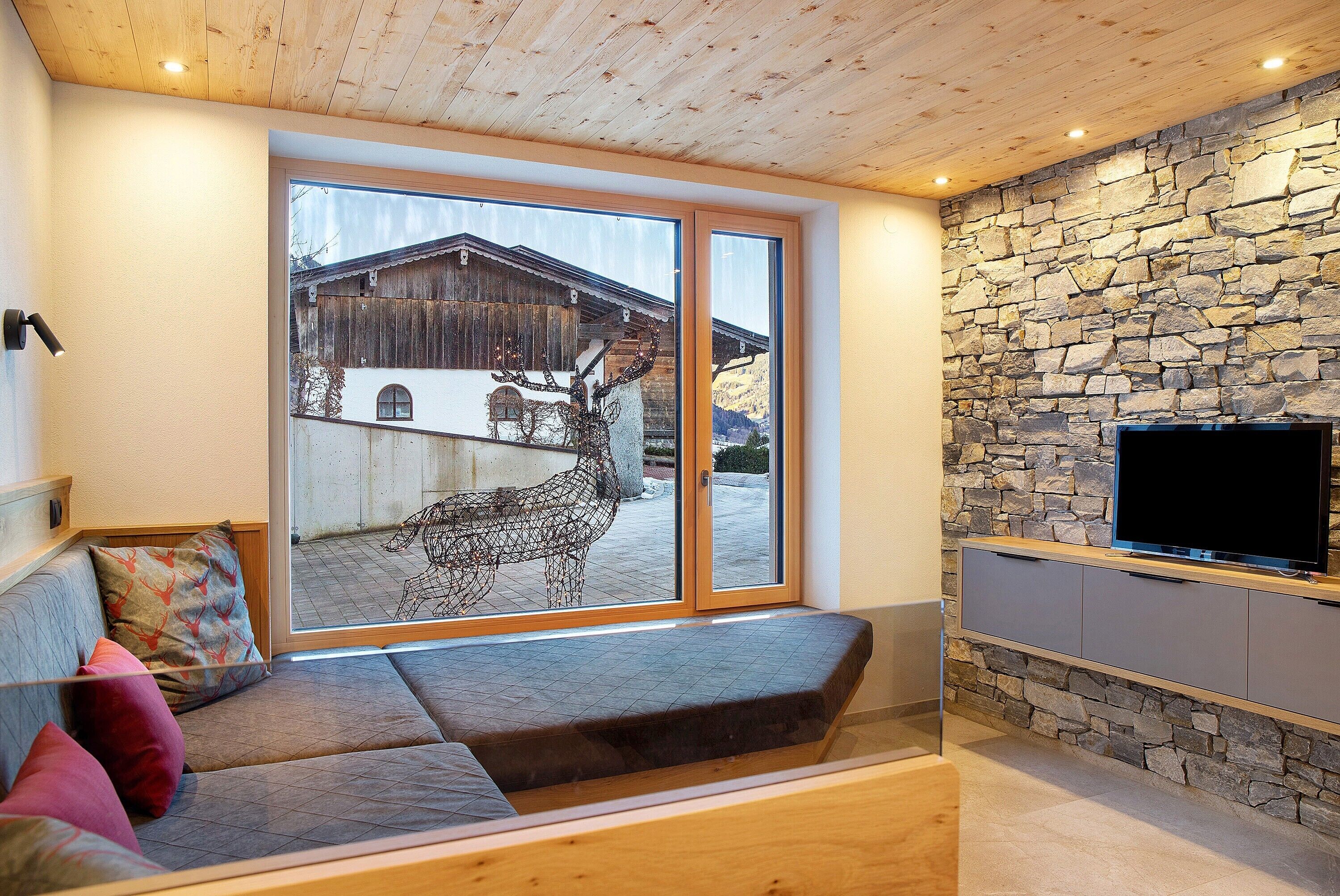 Een moderne woonkamer met een stenen muur, een groot raam met uitzicht op een berg, een houten plafond en een flatscreen tv aan de wand.