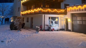 A two-story building with a balcony decorated with lights, a garage, and a snow-covered ground. - Apartment "Wildschön" with Mountain Views, Shared Garden and Wi-Fi (Grafenweg)