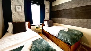 A hotel room with two beds, a wooden headboard, a small table with a lamp, and a window with curtains. - Canterbury Charm - Spacious 4 Bedroom House (Canterbury)