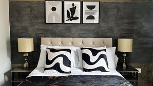 A bedroom with a tufted headboard, two bedside lamps, a bed with a black and white patterned duvet, and three framed artworks on the wall. - Canterbury Charm - Spacious 4 Bedroom House (Canterbury)