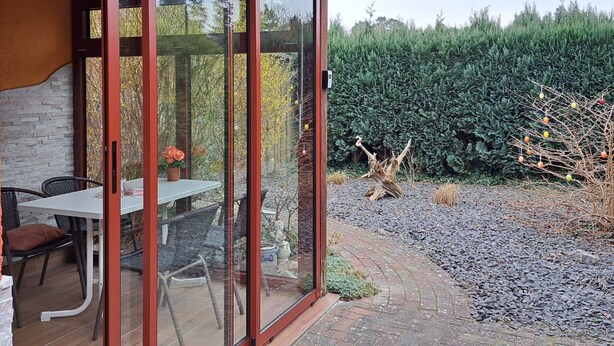 Un patio con tavolo e sedie, un muro in pietra e una porta vetrata che dà su un giardino con siepe e un albero.