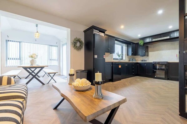A modern kitchen with black cabinets, a wooden dining table, and a view of the living area.