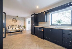 A modern kitchen with black cabinets, a wooden herringbone floor, a large window with a view, and a clock on the wall.