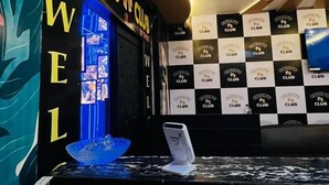 Uma área de bar com um monitor exibindo um gráfico, um telefone em um suporte e uma parede com os dizeres 'CHEER'S CLUB' e 'WELL'.