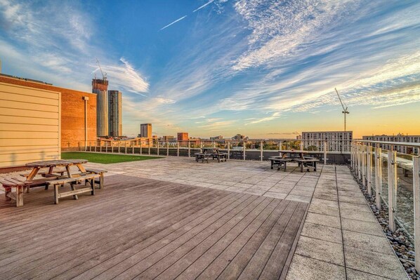 Un patio en la azotea con mesas y bancos de madera, con vistas a un paisaje urbano con edificios y cielo despejado.