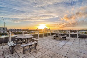 Una terraza en la azotea con mesas de picnic y vistas a la puesta de sol sobre el horizonte de una ciudad.