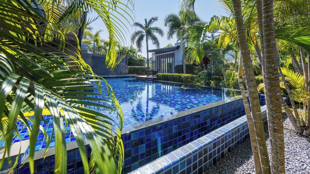 Piscina com azulejos de mosaico azul, cercada por palmeiras e um edifício moderno ao fundo.