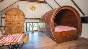 Une chambre à coucher aménagée dans un espace de type « loft », avec un lit en forme de tonneau en bois, une armoire en bois et une table à manger recouverte d’une nappe à carreaux.