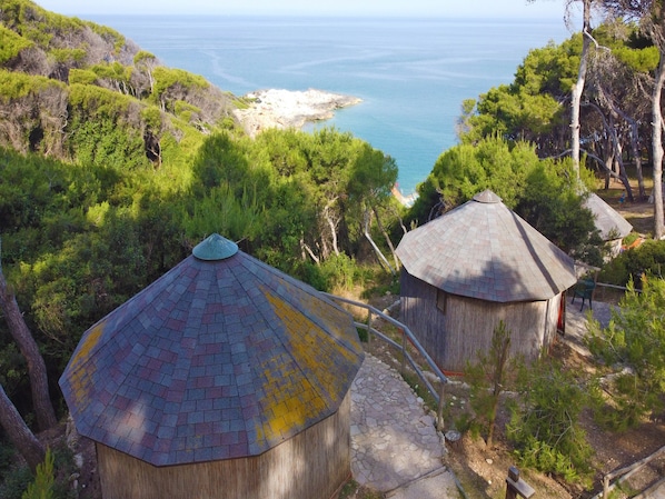 A view of a coastal area with two thatched-roof huts, surrounded by dense greenery and overlooking the sea.