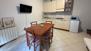 Een compacte keuken met witte kastjes, een magnetron en een kleine koelkast. Er staat een eettafel met stoelen en aan de muur hangt een lijst.