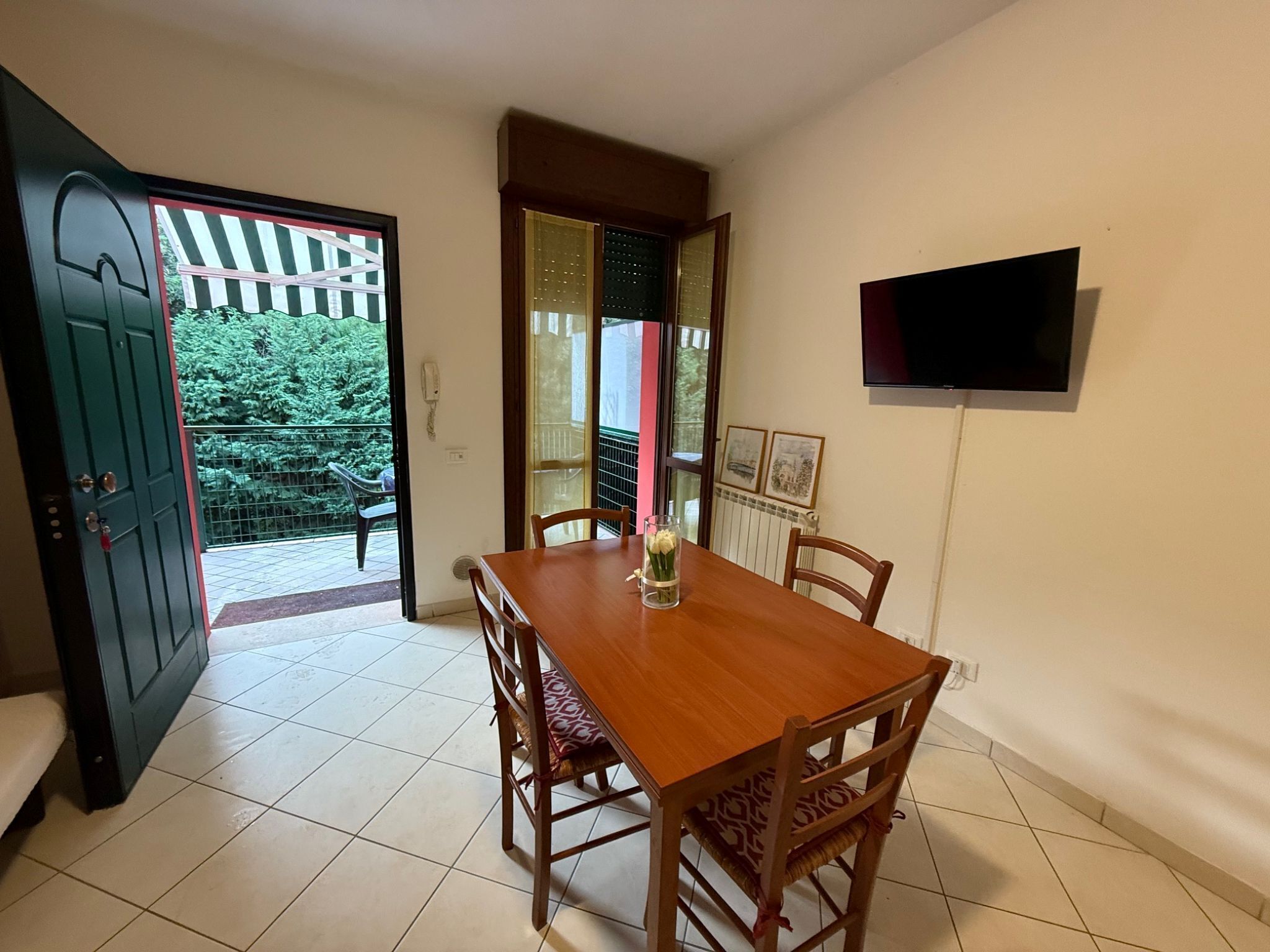 Un comedor con una mesa y sillas de madera, un jarrón pequeño con flores, un televisor montado en la pared y un balcón con vistas a zonas verdes.