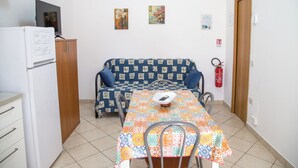 Ett litet pentry med ett kylskåp, ett träskåp och ett bord med en färgglad duk. Det finns en soffa med ett mönstrat överdrag och en brandsläckare som är monterad på väggen.