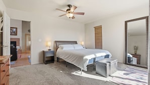 Un dormitorio con cama, lámparas de noche, ventilador de techo, un espejo grande y una pequeña banca.