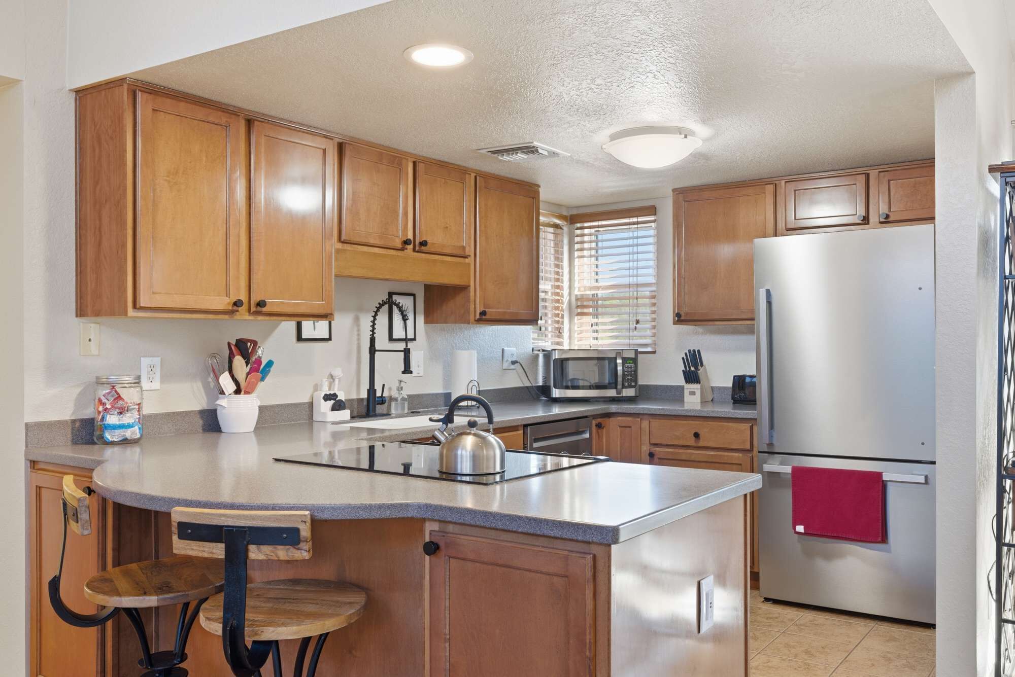 A kitchen with wooden cabinets, a stainless steel refrigerator, a microwave, and a kettle on the island.