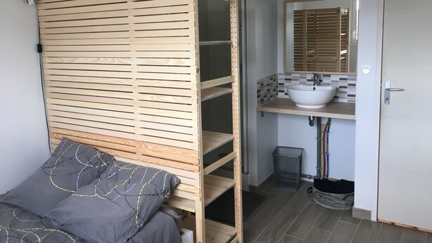 A bedroom with a wooden headboard, a bed with grey bedding, a bathroom with a white sink and tiled backsplash, and a door with a handle. - 1st floor apartment 5 min from town center and 15 min from beaches. (Lannion)