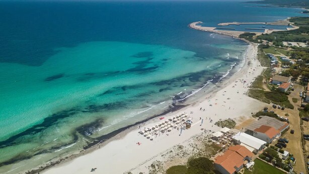 Ett flygbildsbild av ett kustområde med en sandstrand, klart blått vatten och en liten småbåtshamn.