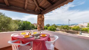 Un espacio comedor al aire libre cubierto, con un pilar de piedra, sillas blancas y una mesa dispuesta con platos, vasos y cubiertos.