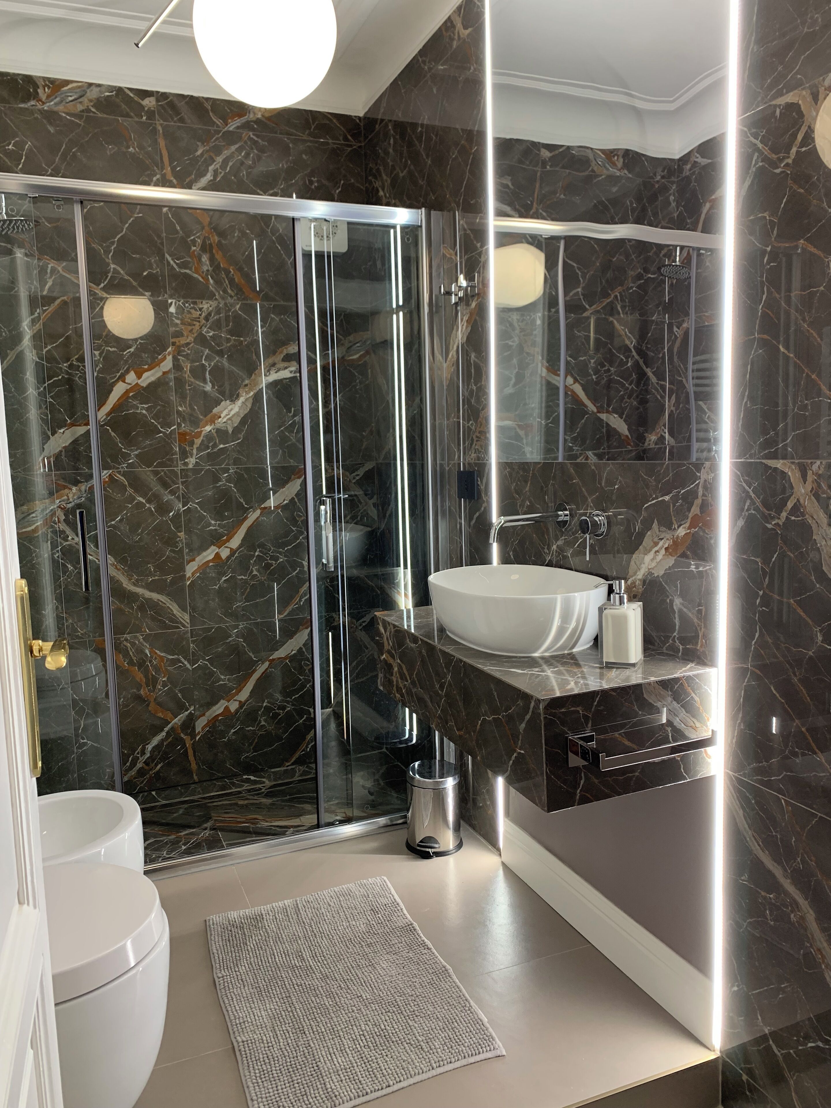 Ein modernes Badezimmer mit einer gläsernen Duschkabine, einem weißen Waschbecken und Marmorwänden.