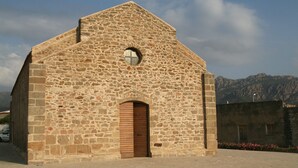 Un edificio de piedra con techo a dos aguas y una puerta central, rodeado de un área pavimentada y montañas al fondo.