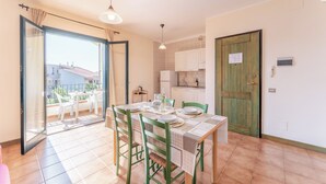 Uma área de refeições com uma mesa posta para quatro pessoas, uma cozinha com armários brancos e uma porta de vidro deslizante que dá para uma varanda externa.