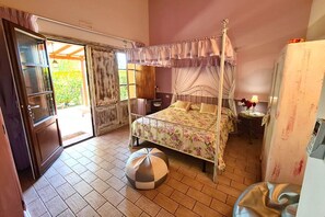 Un dormitorio con cama con dosel, una mesita de noche con lámpara, una pequeña mesa redonda con un florero y un refrigerador.