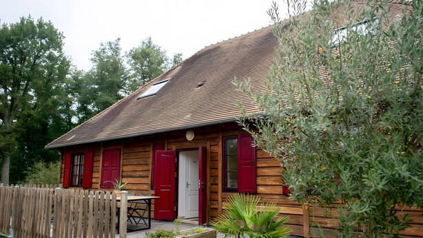 Een houten vakantiehuisje met een rode deur, een klein terras met een tafel en stoelen, en een houten omheining.