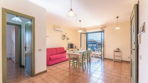 A spacious living area with a red sofa, dining table, and chairs, a kitchen area with a fridge and microwave, and a balcony with a view. - Charming house near Rena Bianca beach (Santa Teresa Gallura)