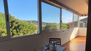 Een balkon met een tafel en stoelen, grote ramen met uitzicht op bomen en gebouwen, en een houten vloer.