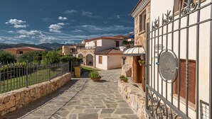 Un patio empedrado con una valla de metal, un pequeño jardín con plantas en macetas y edificios tradicionales con tejados de tejas.