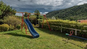 Un parque infantil con un tobogán y columpios en un área verde con una colina al fondo.