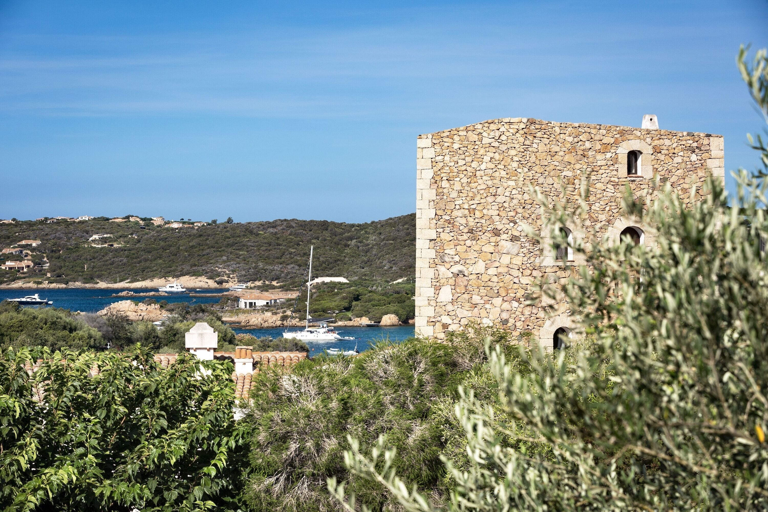 A stone tower with arched windows, surrounded by greenery and overlooking a coastal area with boats.